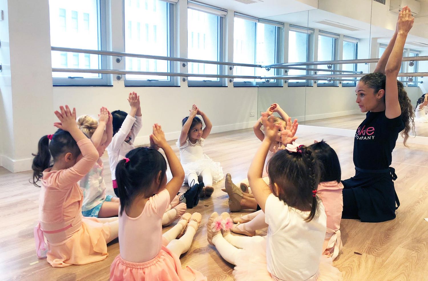 eight little kids and a teacher with their hands in the air in a ballet class